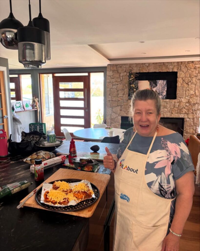 Lady cooking in the kitchen, looking at camera with thumb up: OutAbout Oz 1:1 Person-centred Programs